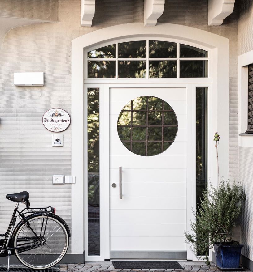 Weißer Hauseingang mit runder Glastür, Topfpflanzen und Fahrrad an grauer Fassade.