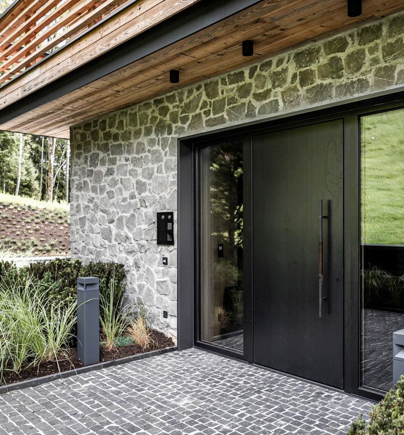 Moderne Eingangstür in dunklem Design mit Natursteinfassade und umliegender Begrünung.